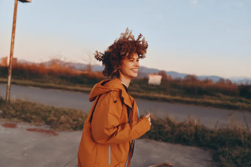 Woman lifestyle outdoors, smiling curly hair in brown backpack, walking along a countryside path at sunset during autumn, warm golden light, soft wind and scenic landscape.