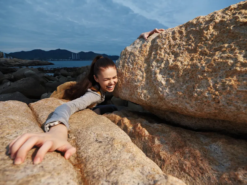 Young woman struggling playfully between large rocks at the beach, embodying joy and adventure with a vibrant coastal background
