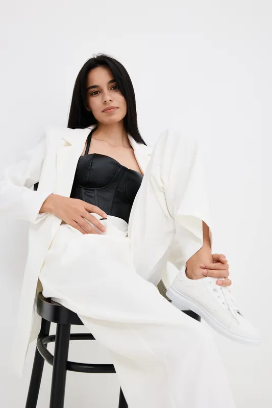 stylish woman wearing white suit and black corset sitting on chair with relaxed confident pose in minimalist studio background, modern fashion portrait