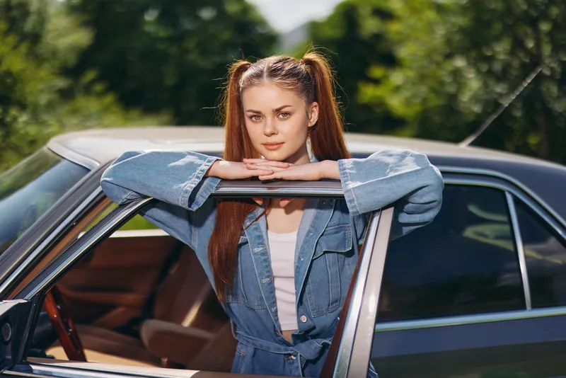 Young woman in denim outfit poses by classic car, exuding confidence and nostalgia in a vibrant green park setting, perfect for lifestyle and automotive themes.