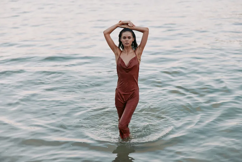 A stylish woman stands waist deep in calm water, wearing a rust red jumpsuit. Her arms raised above her head create poised, confident elegance.