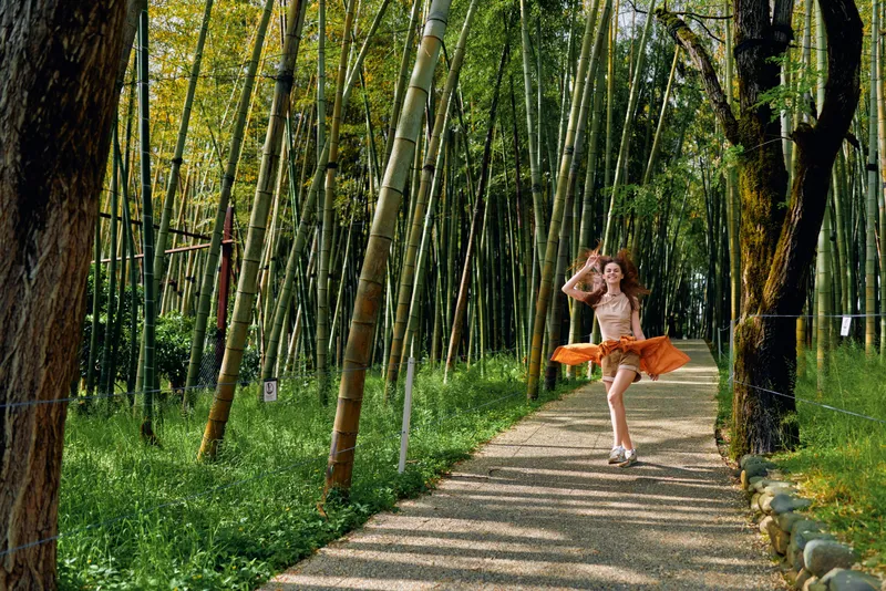 Woman dancer dance in a bamboo forest path wearing an orange skirt, joyful motion and playful pose on a gravel walkway, natural light, serene green trees and park atmosphere