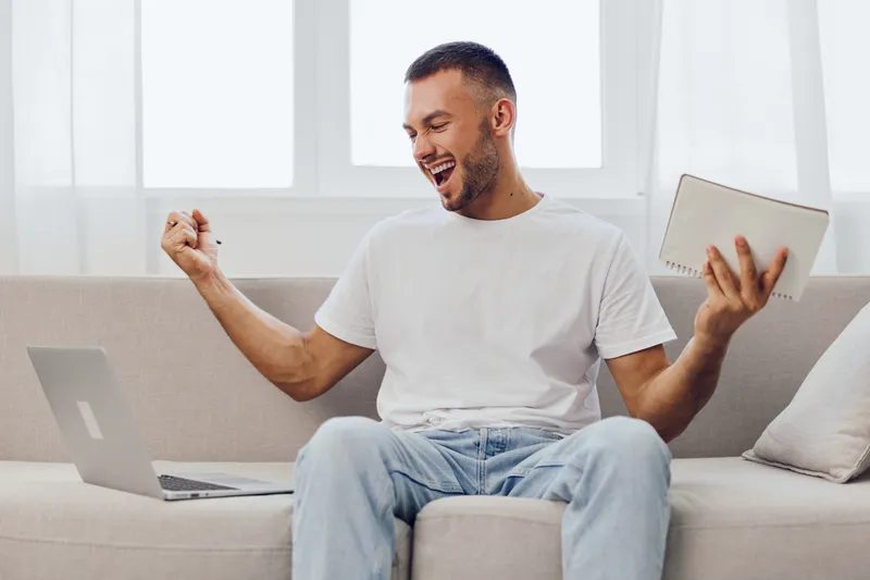 Excited man celebrating success while working on a laptop at home, showcasing modern lifestyle and digital engagement. Perfect for technological themes in contemporary settings.