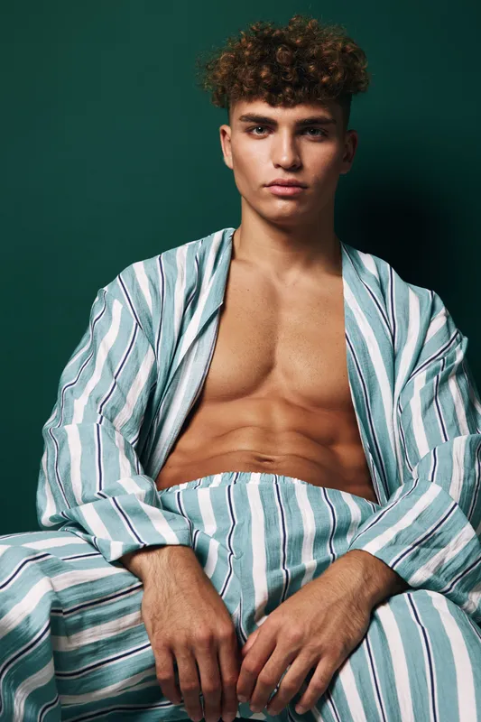 Young man with curly hair wearing striped robe sitting against dark background with serious expression and visible toned chest and abs.