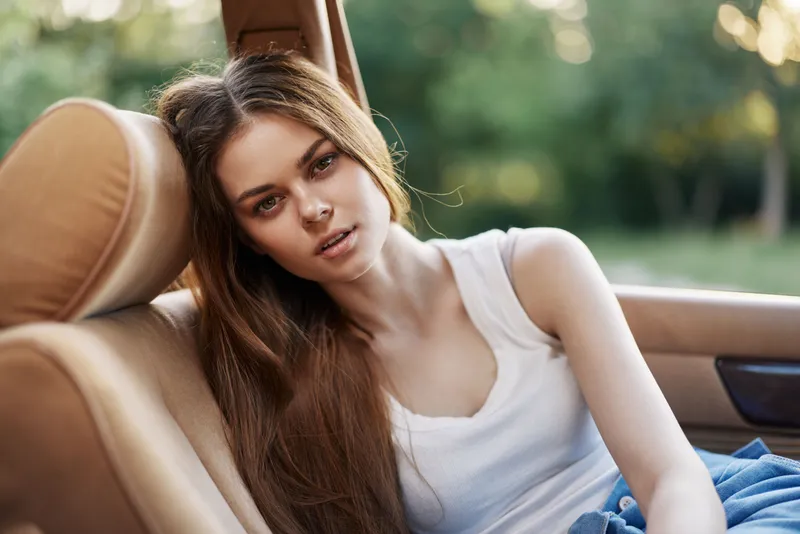 Young woman relaxing in a car, showcasing natural beauty and casual fashion in a serene outdoor setting, with long hair and a laid-back expression.