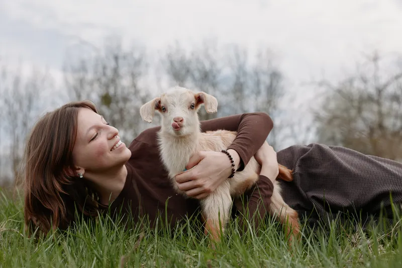Smiling woman lying on grass in an open field, cuddling a small white goat kid. Lifestyle moment focused on connection with animals, nature, and gentle care.