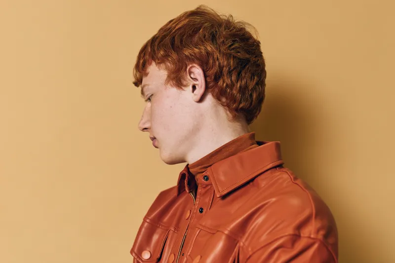 Redhead man in orange leather jacket looking down with neutral expression against beige background. Side profile portrait showing hairstyle and casual fashion details in studio lighting.