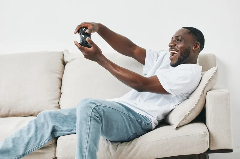 Joyful young African American man playing video games on the couch, wearing a casual white t shirt and jeans, exhibiting excitement and happiness against a neutral background