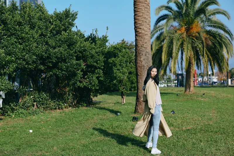 Woman in a long beige coat, jeans and sneakers strolls through a sunny park with palm trees, green grass and a relaxed mood, showcasing outdoor fashion