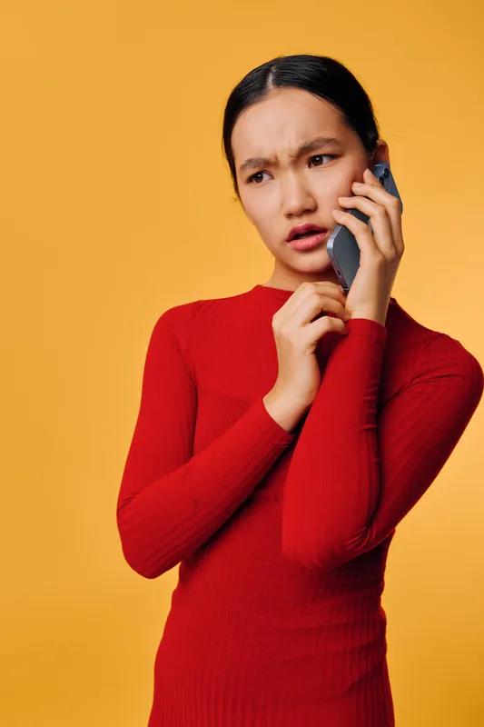 Young woman in red sweater holding phone to ear with concerned expression on orange background, communication and emotion concept, indoor portrait for technology or conversation.