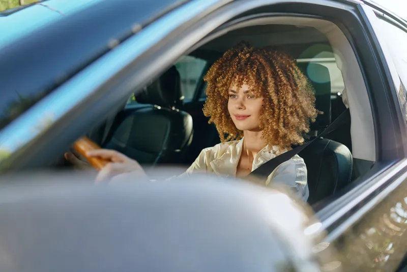 Young woman with curly hair driving a car wearing seatbelt, focused and calm, sunlight illuminating her face, casual white shirt, modern interior.