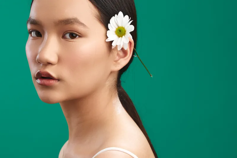 asian woman with a daisy tucked behind her ear gazes softly at the camera against a bright green backdrop, presenting natural beauty and calm confidence.