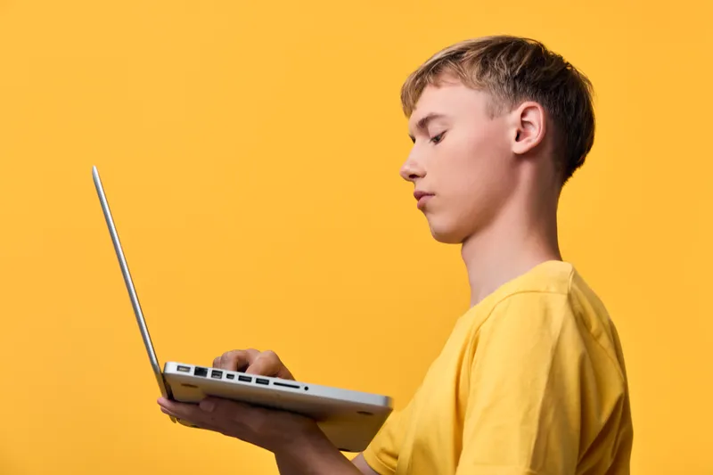 Young learner using a laptop for study and coding on a bright yellow background, focused on screen, keyboard and device, education, digital skills, remote learning, creativity
