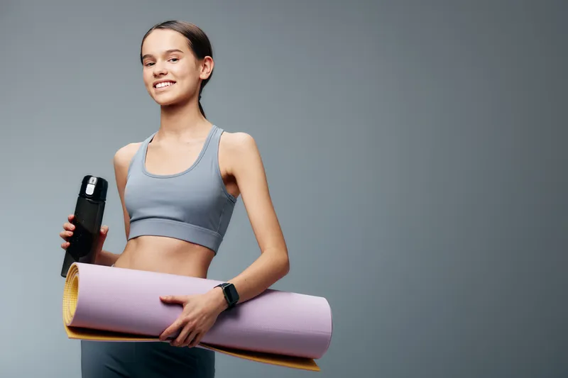 Fit young woman in athletic wear holds a yoga mat and water bottle against a neutral studio backdrop, ready for a workout session and healthy lifestyle.