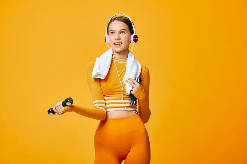 Woman in bright orange athleticwear holding a dumbbell, with a towel over her shoulder and headphones, posing for a dynamic workout scene in a bold studio.