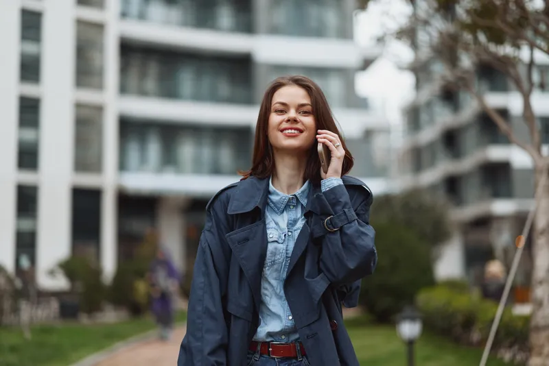 Young woman in denim shirt and navy jacket stands outdoors in an urban courtyard, smiling confidently as she adjusts her hair, ready for a casual city day