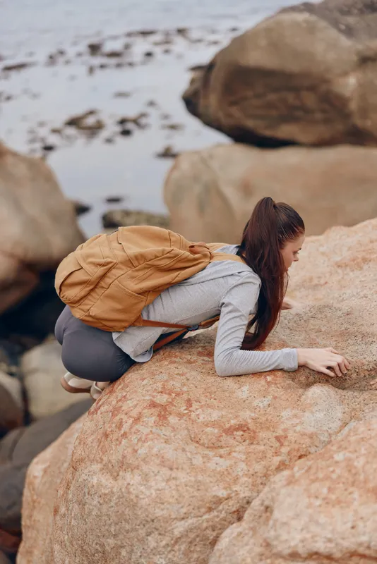Young woman climbing on rocky shore, wearing casual gray clothing and a brown backpack, showcasing determination and adventure against a coastal background