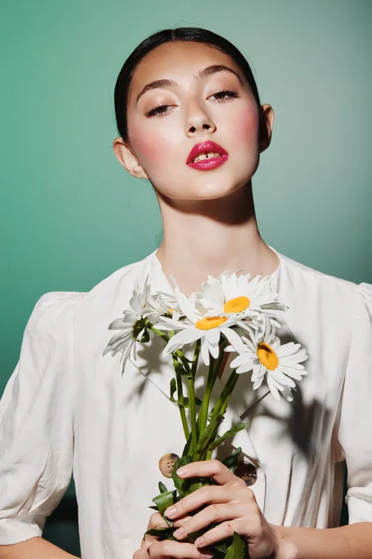 Young woman with sleek hair holds white daisies wearing soft puffed sleeve dress. Stylish bright makeup with glossy pink lips. Fashion and beauty concept with minimalist background.