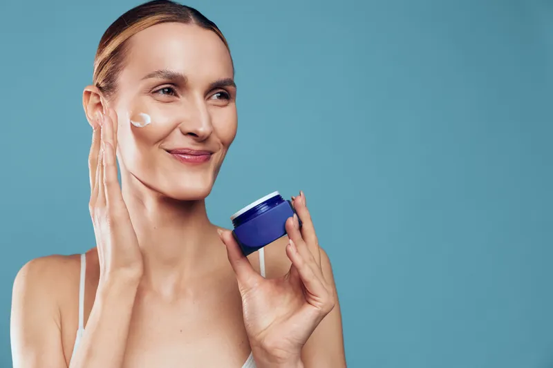 Woman skincare routine, beauty model applies face cream and moisturizer, holding a jar, smiling, representing hygiene and dermatology, isolated on plain blue studio background for self care