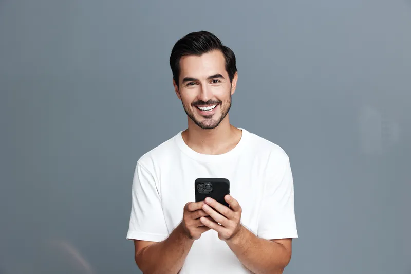 Smiling man in casual white t-shirt holding smartphone while looking at the camera with a joyful expression against a plain grey background. People lifestyle concept.