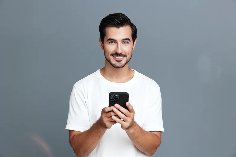 Smiling young man in a white t-shirt joyfully holding a smartphone, looking at the camera, with a neutral gray background creating a modern lifestyle vibe.