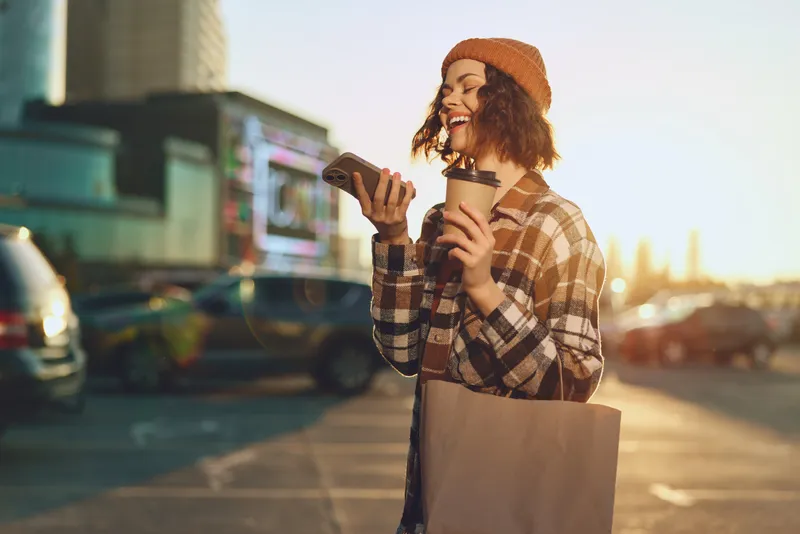 Woman holding coffee and phone with a bright smile on an urban street at golden hour sunlight, beanie and tote in a candid scene of authenticity, mindful living and emotional storytelling.
