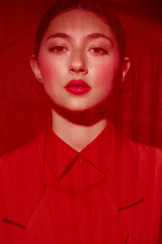 Portrait of young woman in vibrant red shirt with glossy red lips behind a red veil. Creative beauty and fashion concept with artistic moody lighting and intense color contrast.