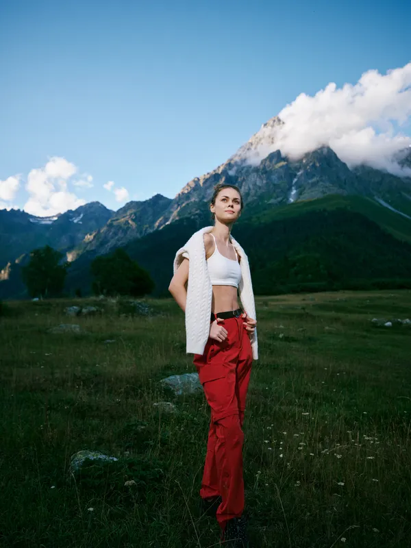 A woman stands in a meadow with distant mountains and a blue sky, wearing a white top and red pants, capturing a calm outdoor lifestyle and adventure vibe.
