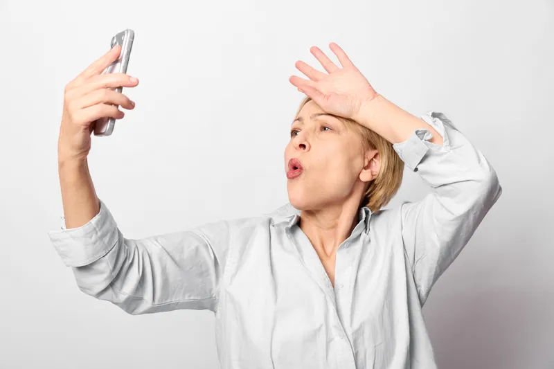 Woman with smartphone, elderly surprised expression, hands up in studio, white background, open mouth shout gesture, communication moment, soft intense light