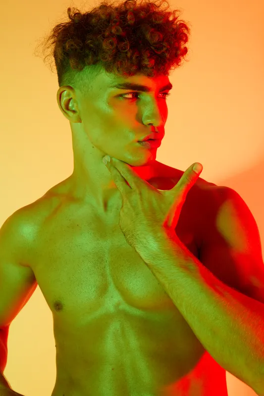 Young shirtless man with curly hair posing with hand on chin and confident facial expression under vibrant red and green lighting against yellow background in studio portrait.