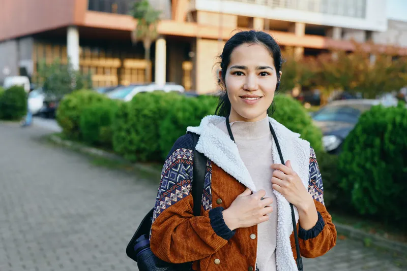 Woman outdoors in a casual street style wearing a coat and backpack, urban setting with greenery and buildings, confident smile and relaxed pose for fashion and lifestyle storytelling