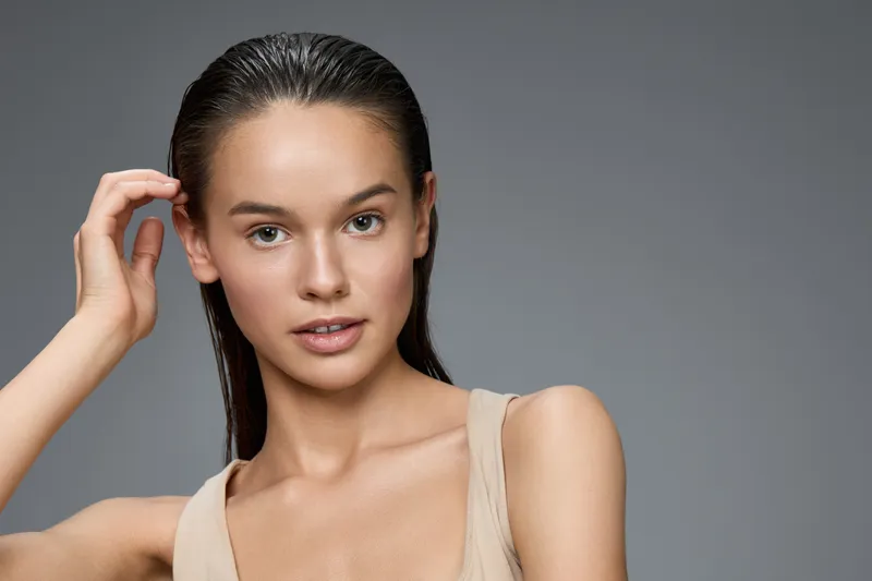 Natural beauty portrait of a young woman with wet hair, showcasing minimal makeup and a neutral outfit against a soft gray background, exuding confidence and freshness