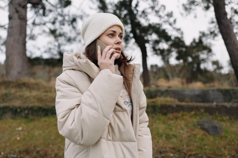 Woman talking on a mobile outside in winter, wearing a beige coat and beanie, in a park with bare trees and a muted landscape distance.