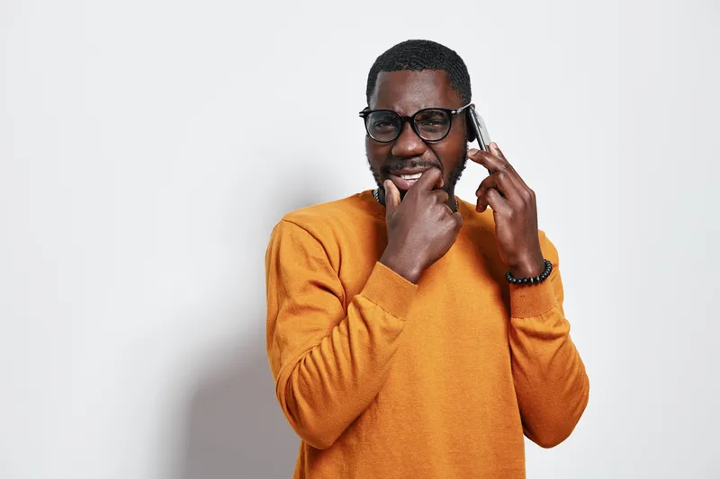 African man with dark skin wearing glasses and orange sweater talking on smartphone with thoughtful expression in studio. Communication, technology and casual style with confident pose.