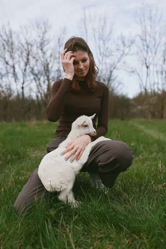 Young woman kneels in green grass gently holding a white baby goat outdoors on a cloudy day with bare trees in the background, showing calm connection and natural lifestyle.