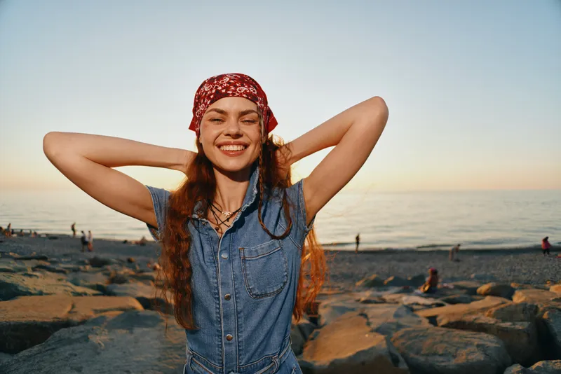 Lifestyle woman in modern boho-western denim street style enjoys sunset by the sea with a red bandana and relaxed, joyful expression in warm film color tones.