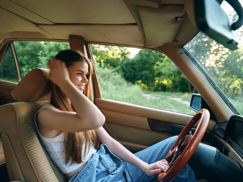 Young woman driving in a vintage car, smiling, wearing casual denim clothing, showcasing a carefree summer vibe with green nature background.