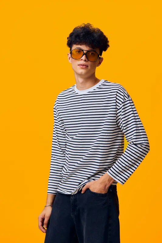 Young man wearing striped shirt and yellow sunglasses posing confidently against bright orange background, showcasing casual style and modern fashion trends.