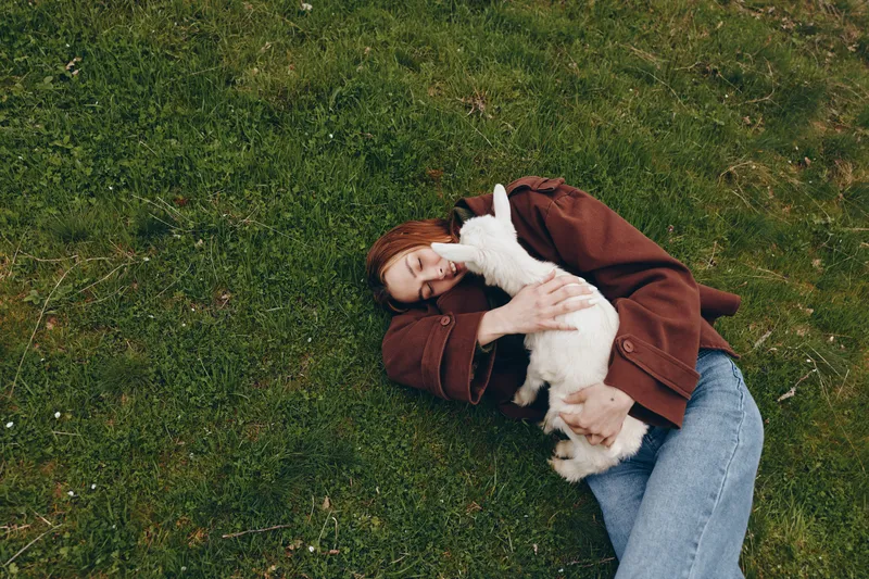 goat, kid, lifestyle, outdoors, affection, cuddle, human, grass, animal, field A person lies on the grass, wearing a brown jacket, smiling softly while embracing a small goat kid in a warm moment