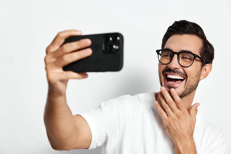 Smiling man with glasses happily taking a selfie while wearing a white t-shirt, showcasing joy and enthusiasm against a solid light background.