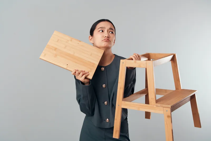 Woman holding wooden chair and board with thoughtful expression for furniture design, craftsmanship, carpentry and creativity in workshop or construction space.