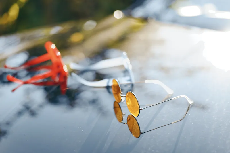 A pair of sunglasses rests on a sleek car hood under bright daylight. The reflective surface and warm glow create a casual, relaxed mood and a modern, stylish outdoor scene.