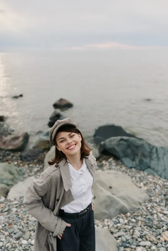 Smiling young woman in casual outfit and hat stands on rocky beach near calm sea under cloudy sky. Concept of outdoor lifestyle, happiness, travel, and nature connection.