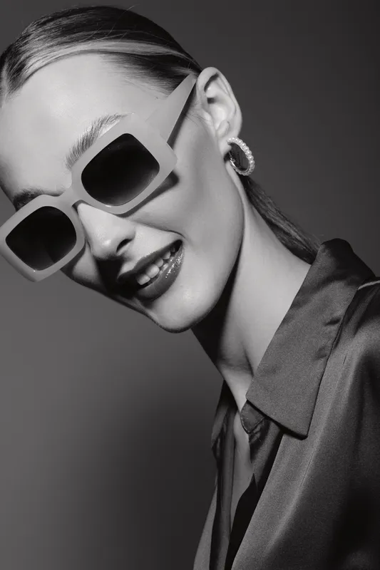 Stylish white woman with pale skin wears large square sunglasses and hoop earrings. She smiles confidently in a smooth satin shirt against a neutral background. Fashion portrait concept.