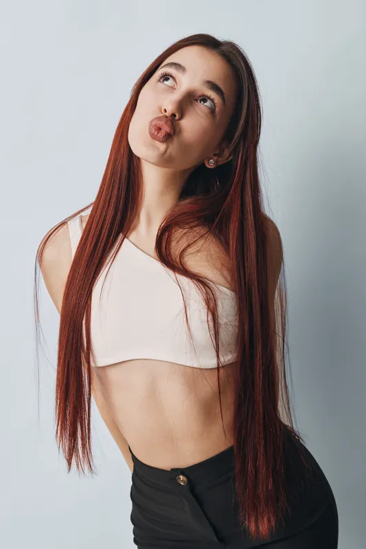 Young woman with long red hair making a playful face with pursed lips standing against a light background. Casual style with white top and black pants in studio portrait.