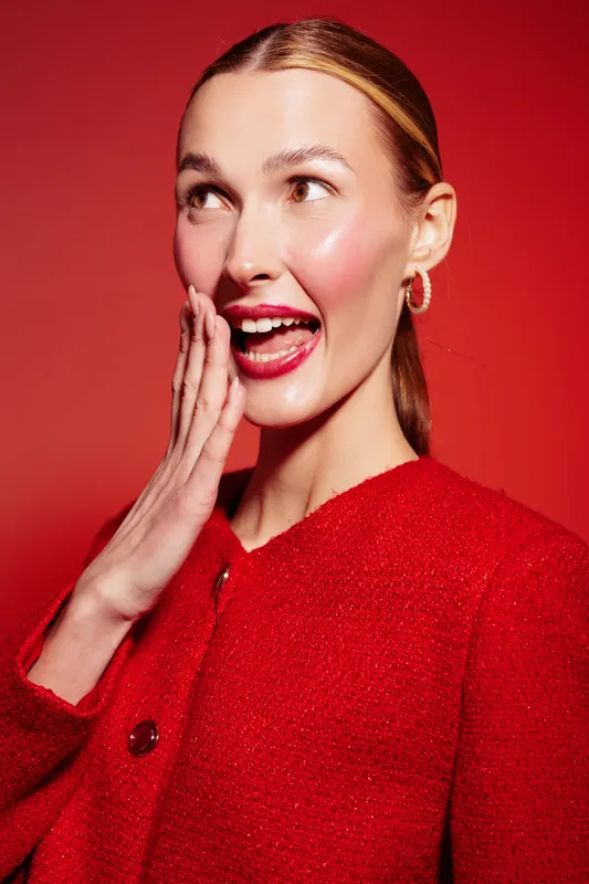 Young woman with shiny skin and red lips wears red jacket and earrings, posing with hand near mouth on red background. Concept of fashion, beauty, glamour, and joyful expression.