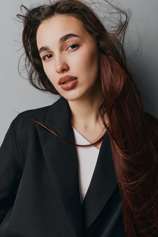 Young woman with long red hair wearing black blazer and white shirt posing against gray background. Portrait of confident female model with natural makeup and loose hairstyle indoors.