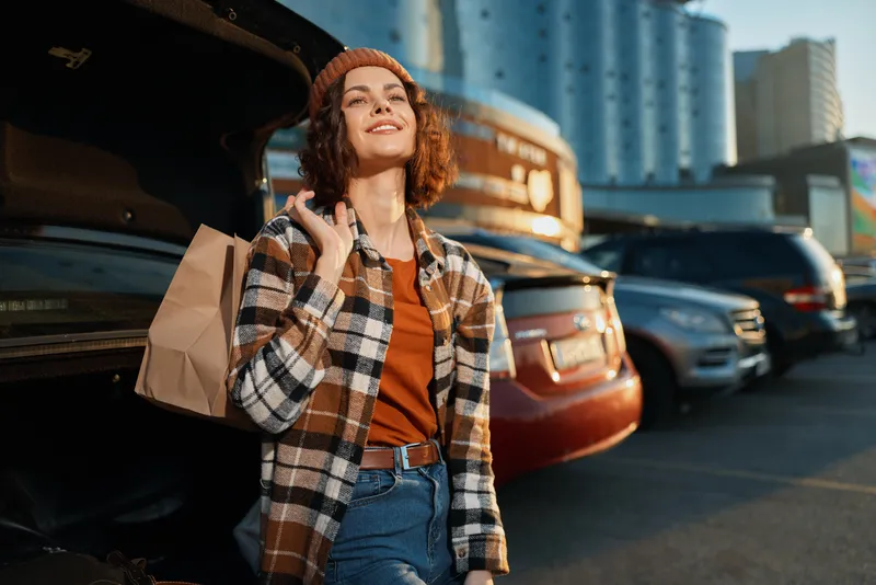 Woman shopping in urban parking with a warm smile, authenticity and candid lifestyle captured at golden hour glow, carrying paper bag by open car trunk for mindful living and emotional storytelling.