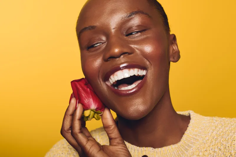 Happy smiling dark skinned African woman with natural makeup holds red flower near face on yellow background. Concept of joy, beauty, natural skin, and floral freshness in studio portrait.