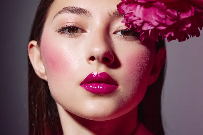 Close-up portrait of a young woman with vibrant pink lipstick and blush adorned with a matching flower. Studio lighting enhances the beauty creative and fashion concept with intense colors.